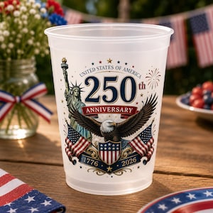 May include: A clear plastic cup with a patriotic design celebrating the 250th anniversary of the United States. The design includes the Statue of Liberty, an eagle, American flags, and the years 1776 and 2026.