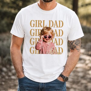 Pu&ograve; includere: T-shirt bianca con la scritta dorata "GIRL DAD". La maglietta presenta una foto di un bambino piccolo con occhiali da sole. Il nome del bambino, "Olivia", &egrave; scritto in corsivo sotto.
