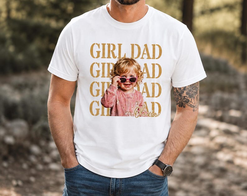 Pu&ograve; includere: T-shirt bianca con la scritta dorata "GIRL DAD". La maglietta presenta una foto di un bambino piccolo con occhiali da sole. Il nome del bambino, "Olivia", &egrave; scritto in corsivo sotto.