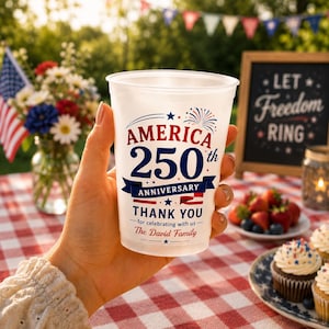 May include: A clear plastic cup with the text "AMERICA 250th ANNIVERSARY THANK YOU" in red and blue. The cup is held in a hand, with a picnic setting in the background, including cupcakes and an American flag.