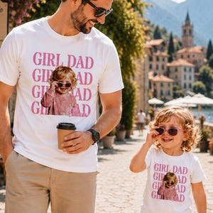 Pu&ograve; includere: T-shirt bianche con la scritta rosa "GIRL DAD" e una foto di una bambina con gli occhiali da sole. Le magliette sono indossate da un uomo e da una bambina. L'uomo tiene in mano una tazza di caff&egrave;.
