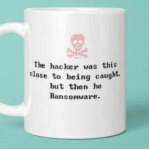 May include: A white ceramic mug featuring a red binary code skull and crossbones graphic above the black text: The hacker was this close to being caught, but then he Ransomware. The mug has a curved handle on the left side and a smooth finish.