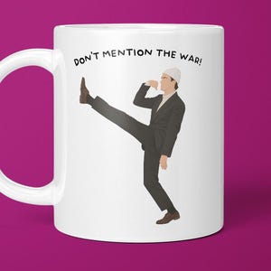 May include: White ceramic mug with a handle featuring the text "DON'T MENTION THE WAR!" and an illustration of a man in a suit kicking. The man is wearing a white hat. The mug is set against a magenta background.