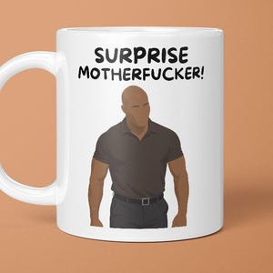 May include: White ceramic mug with the text "SURPRISE MOTHERFUCKER!" in black. The mug features an illustration of a man in a black polo shirt and black trousers. The mug has a white handle and is set against a brown background.