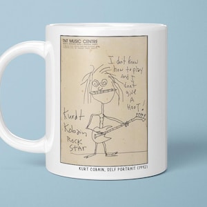 May include: White ceramic mug with a cartoon drawing of Kurt Cobain playing guitar. The drawing includes the text "I don't know how to play and I don't give a hoot!" and "Kurt Kobain Rock Star".