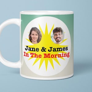 May include: White ceramic mug with a handle, featuring a design with two faces, a yellow burst, and the text "Jane & James In The Morning" in black and red. The mug is set against a light blue background.