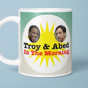 May include: White ceramic mug with the text "Troy & Abed In The Morning" and images of two men. The mug has a white handle and a light blue background. The design includes a yellow burst.