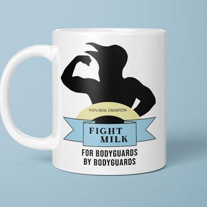 May include: White ceramic mug with a black silhouette of a muscular figure flexing. The mug features the text "FIGHT MILK" in a blue banner, with additional text below. The mug is on a light blue background.