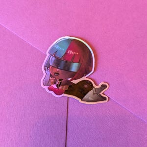 May include: A holographic sticker featuring a cartoon character with a helmet-like visor. The character is depicted in a reclining pose, with a color scheme of red, blue, black, and gray. The sticker is placed on a pink surface.