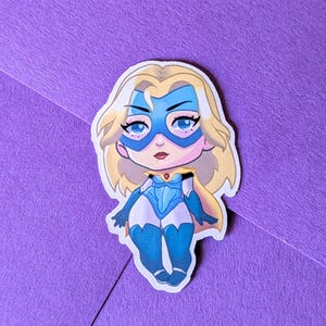 May include: A sticker featuring a cartoon superheroine with blonde hair, a blue mask, and a blue and white costume. The sticker is on a purple background.