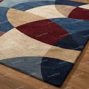 May include: A rectangular rug featuring a geometric design of overlapping arcs in blue, beige, and burgundy. The rug is made of a soft, textured material. It is placed on a wooden floor. The rug's design is modern.