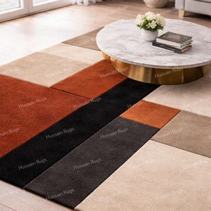 May include: Geometric patterned area rug in shades of beige, brown, black, and rust. The rug is placed on a hardwood floor, with a marble-topped coffee table and a window in the background. The rug has the text "Hussain Rugs" repeated across it.