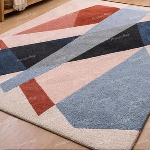 May include: Geometric patterned area rug in shades of blue, coral, gray, and cream. The rug features a modern design with angular shapes and a textured surface. The rug is rectangular and appears to be made of a soft material.