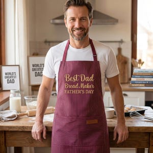 May include: A burgundy and white striped apron with the text "Best Dad Bread Maker FATHER'S DAY" in gold script. The apron has a front pocket and is worn by a person in a kitchen setting.