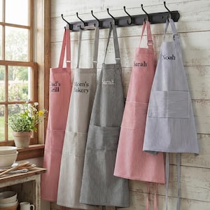 May include: Five striped aprons hanging on a black wall-mounted rack with hooks. The aprons feature various colours including red, grey, black, and blue, with embroidered text including Dad's Grill, Mom's Bakery, Sarah, and Noah on the front.