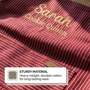 May include: A red and white pinstriped apron featuring gold embroidered text that reads Sarah Cookie Queen. The apron includes a front pocket with a small brown rectangular tag. A white informational box is overlaid with the text STURDY MATERIAL.
