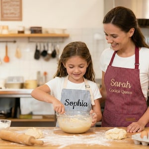 May include: A child wearing a blue and white pinstriped apron with the name Sophia embroidered in black, and an adult wearing a maroon and white pinstriped apron with the name Sarah and the text Cookie Queen embroidered in gold thread.