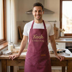 May include: A burgundy apron with thin white vertical stripes, featuring the embroidered text "Noah" and "Cookie King" in gold. The apron includes a large front pocket with a small rectangular brown leather-like patch on the upper right corner.