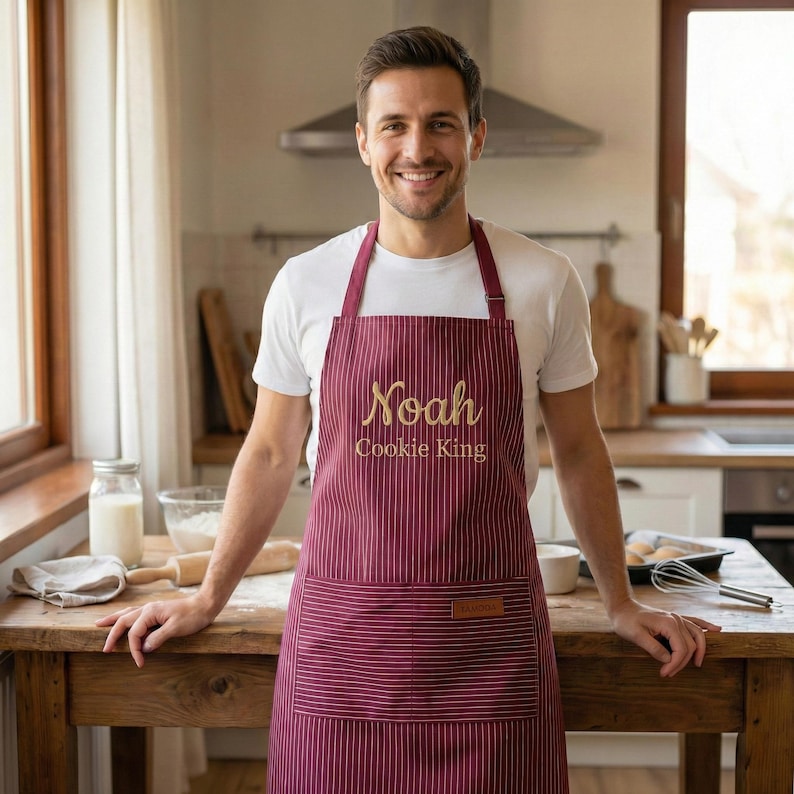 May include: A burgundy apron with thin white vertical stripes, featuring the embroidered text "Noah" and "Cookie King" in gold. The apron includes a large front pocket with a small rectangular brown leather-like patch on the upper right corner.