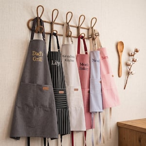 Può includere: Assortimento di grembiuli a righe appesi a un supporto in legno. Ogni grembiule presenta un colore e un testo diversi, tra cui "Dad's Grill", "Lily", "Grandma's Kitchen", "Mom's Bakery", "Noah" e "Sara". Sono visibili anche un cucchiaio di legno e steli di cotone.