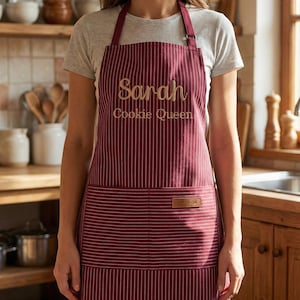 May include: A burgundy apron with thin white vertical stripes, featuring the embroidered text "Sarah" and "Cookie Queen" in gold thread on the chest, and a small rectangular brown leather-like patch on the front pocket area.