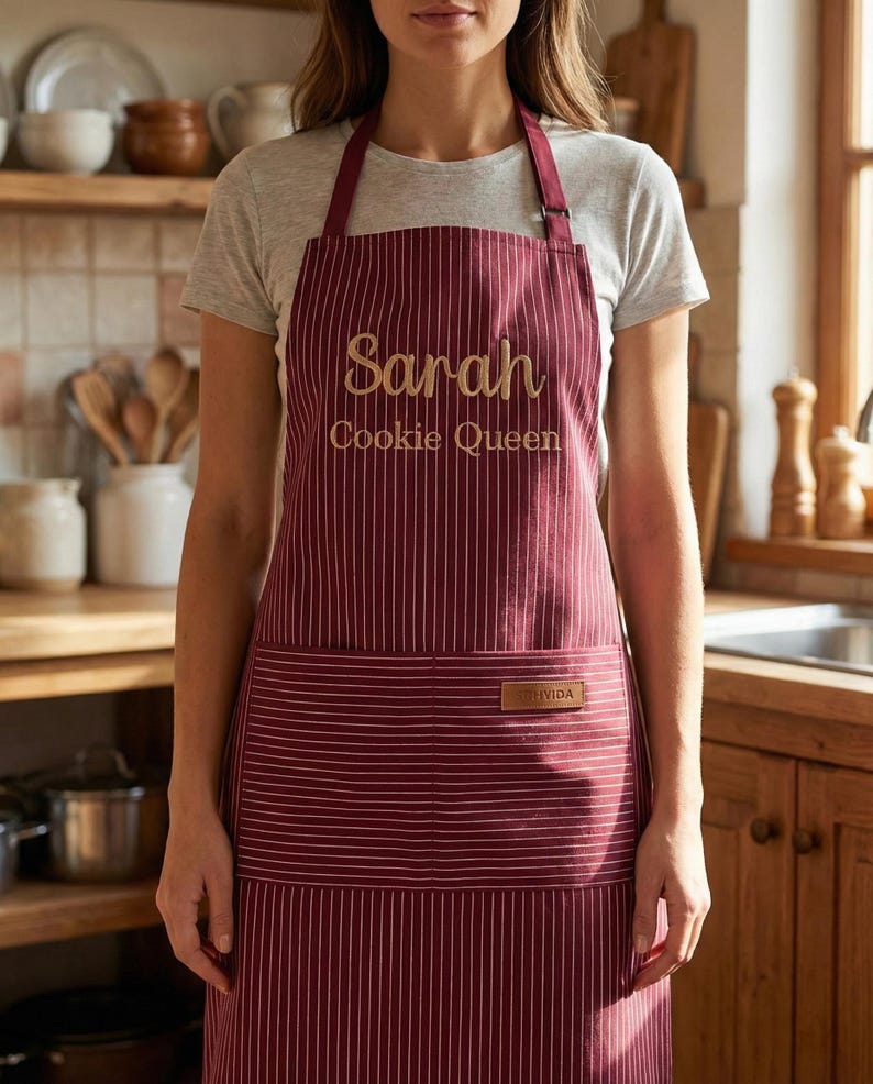 May include: A burgundy apron with thin white vertical stripes, featuring the embroidered text "Sarah" and "Cookie Queen" in gold thread on the chest, and a small rectangular brown leather-like patch on the front pocket area.