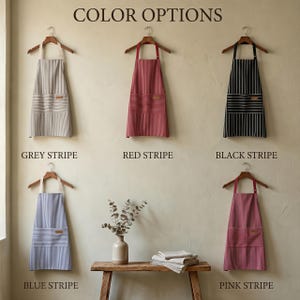May include: Five aprons hanging on wooden hangers, labelled Colour Options. The aprons feature vertical stripes in grey, red, black, blue, and pink. Each apron has a front pocket with horizontal stripes and a small rectangular patch on the pocket.