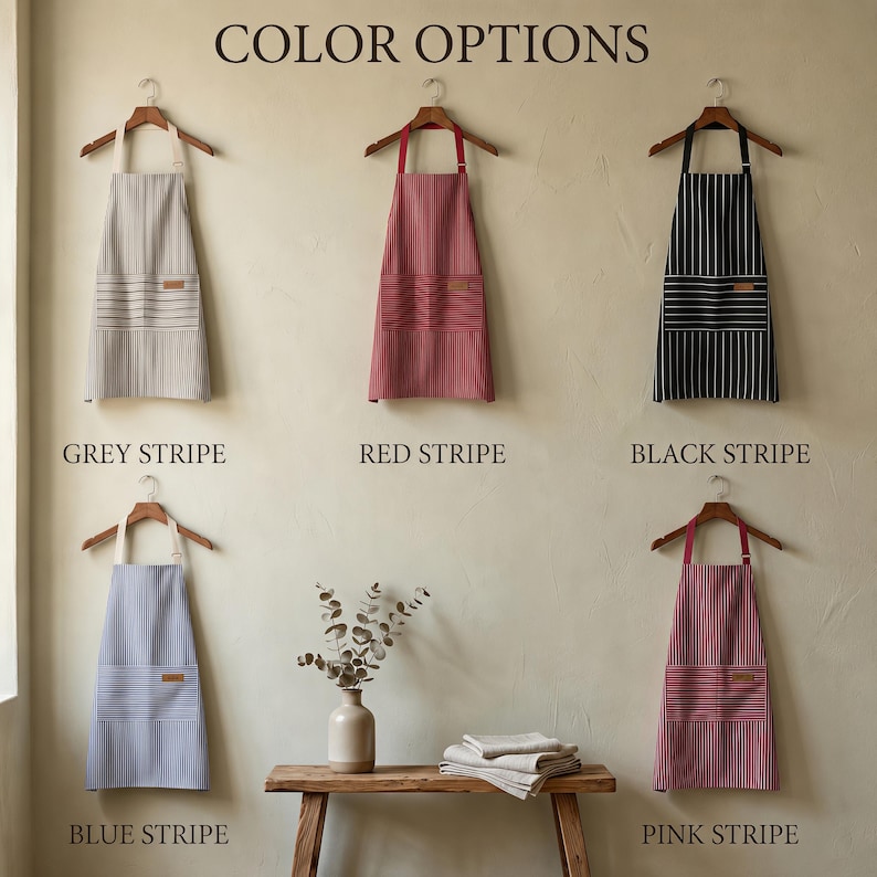 May include: Five aprons hanging on wooden hangers, labelled Colour Options. The aprons feature vertical stripes in grey, red, black, blue, and pink. Each apron has a front pocket with horizontal stripes and a small rectangular patch on the pocket.