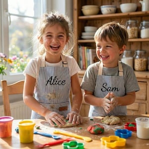 May include: Two children wearing matching blue and white striped aprons with the names Alice and Sam embroidered in black script. They are standing at a wooden table covered in colourful modelling clay, various plastic tools, and cutters.