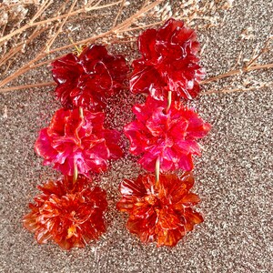 May include: A pair of handmade earrings featuring three tiers of flower-shaped pendants in shades of red and orange. The earrings are suspended from gold-colored hooks. The background is a glittery, neutral color.