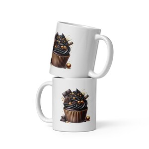 Puede incluir: Dos tazas de cerámica blanca apiladas una encima de la otra. Cada taza presenta una ilustración detallada de un cupcake con glaseado oscuro, detalles de chocolate y chispas doradas. Los cupcakes están sobre un fondo blanco.