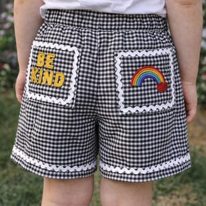 Puede incluir: Shorts de cuadros blancos y negros con ribete festoneado blanco. Los shorts tienen dos bolsillos, uno con el texto bordado "BE KIND" en amarillo y otro con un diseño de arcoíris y corazón.