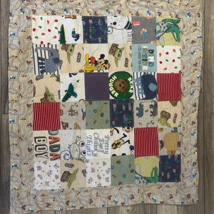 May include: A patchwork baby quilt with squares of fabric featuring cartoon characters, stripes, and text. The quilt has a beige border with a road and car pattern. The squares include the text "DADA BOY" and "Pretty Eyes Chunky Thighs".