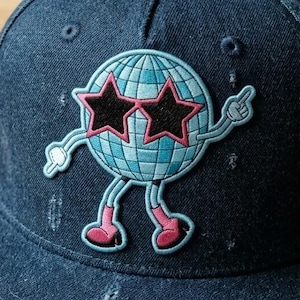 May include: A dark blue denim baseball cap featuring a cartoon disco ball character with star-shaped sunglasses and pink boots. The cap has a curved brim and a button on top. Sunglasses are visible in the background.