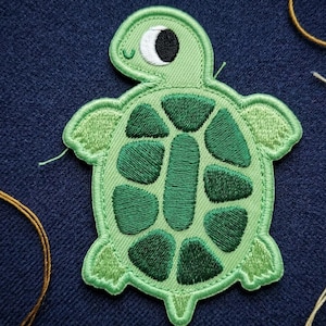 May include: An embroidered turtle patch featuring a light green body and a darker green shell with distinct segmented patterns. The turtle has a white eye with a black pupil and a small stitched smile, rendered in a flat, two-dimensional style.