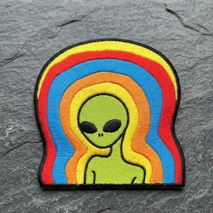 May include: Embroidered patch featuring a green alien with black eyes and a smiling mouth. The alien is set against a rainbow-colored background of red, orange, yellow, and blue. The patch has a black border.