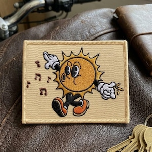 May include: Embroidered patch featuring a cartoon sun with a smiling face, black shorts, and orange shoes. The sun is surrounded by yellow rays and musical notes. The patch has a tan border and is on a brown leather surface.