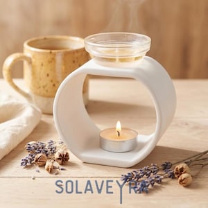 May include: A white ceramic essential oil diffuser with a lit tea light candle. A small glass bowl holds oil, with steam rising. A speckled mug and lavender sprigs are nearby. The word "SOLAVEYRA" is at the bottom.