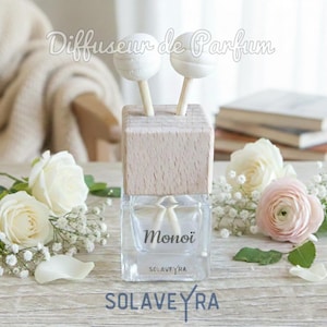 May include: A clear glass diffuser bottle with a wooden top and two white wooden ball sticks. The bottle has the word "Monoï" printed on it, and is surrounded by white roses and baby's breath. The brand name "SOLAVEYRA" is printed on the bottle.