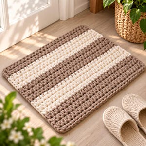 May include: A rectangular floor mat featuring a thick, chunky crochet texture with alternating horizontal stripes in shades of cream and light brown. The mat has a soft, knotted appearance with a consistent weave pattern throughout the entire surface.
