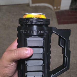 Może przedstawiać: Czarny "Assault Koozie" z uchwytem, trzymający puszkę z żółtymi i pomarańczowymi akcentami. Koozie ma teksturowany design ze słowami "ASSAULT KOOZIE" wydrukowanymi z boku. Koozie jest przeznaczony do przechowywania puszki o standardowym rozmiarze.