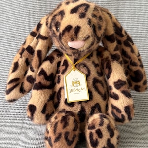 Pode incluir: Um coelho de pelúcia com estampa de leopardo com orelhas compridas, nariz rosa e uma etiqueta que diz "Jellycat London". O bicho de pelúcia é marrom e bege com manchas marrons escuras. O coelho está em uma superfície cinza texturizada.