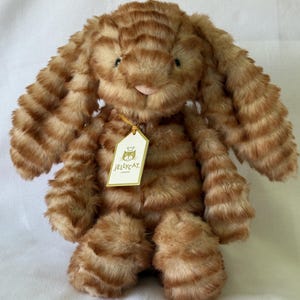Puede incluir: Un conejo de peluche a rayas naranja y marrón con largas orejas caídas. El peluche tiene una pequeña nariz rosa y una etiqueta que dice "Jellycat London."