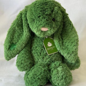 Puede incluir: Un peluche verde con forma de conejo, con orejas largas y caídas y un aspecto suave y texturizado. El conejo tiene una nariz rosa y un pequeño detalle de trébol en una pata. Una etiqueta verde con el logo "Jellycat" está adjunta.