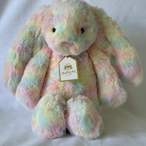 Puede incluir: Un conejito de peluche de color arcoíris pastel con largas orejas caídas. El peluche tiene una textura suave y esponjosa y una etiqueta que dice "JELLYCAT. LONDON."