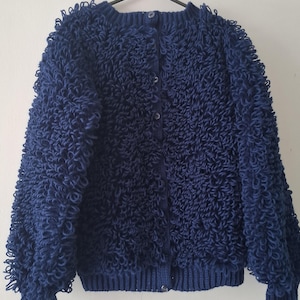 May include: A dark blue long-sleeve cardigan featuring a textured loop-stitch knit pattern throughout the body and sleeves. The garment includes a ribbed crew neckline, a ribbed hem, and a vertical front placket secured with five matching buttons.