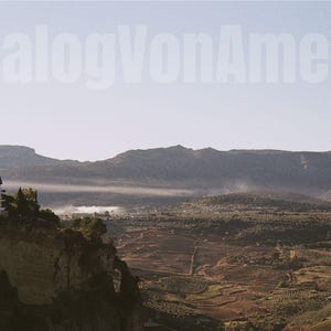 Può includere: Fotografia di paesaggio con un edificio su una scogliera e il testo "AnalogVonAmelie" nel cielo. L'immagine cattura una catena montuosa, una valle e un cielo sereno, evocando tranquillità.