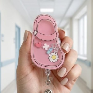 May include: A pink badge reel in the shape of a clog, featuring medical-themed graphics like a heart, cross, stethoscope, pill, and flower. The reel has a clear plastic holder and a retractable cord.