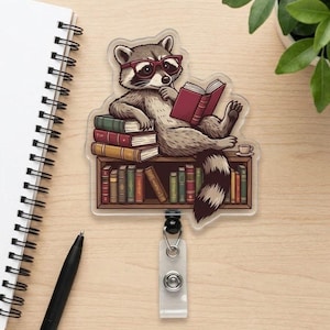 May include: A clear, rectangular badge reel featuring a cartoon raccoon wearing glasses and reading a book while sitting on a stack of books. The raccoon is brown and gray with a striped tail. A small teacup is next to the raccoon.
