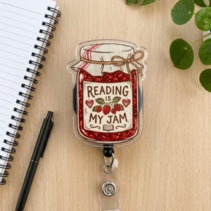 May include: A clear acrylic badge reel with a graphic of a jam jar. The jar is filled with red jam and has the text "Reading is my jam". The reel has a retractable cord and a clear plastic clip.